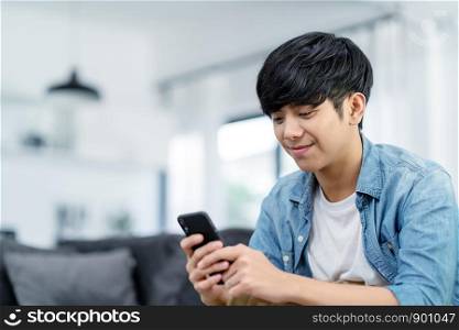 Happy asian teenager using smart phone and smiling on sofa living room at home. Asian man holding and using cellphone for searching data and social medie on internet. Teenager and Technology concept.