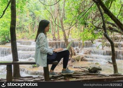 Happy Asian woman, a freelance, working and using a computer laptop notebook at waterfall in the tropical forest during travel trip and holidays vacation outdoors at national park, Thailand.