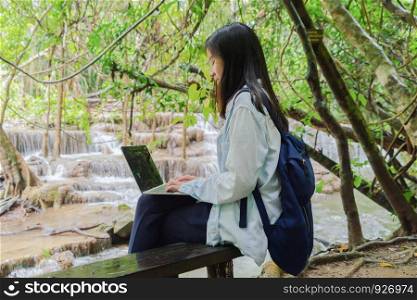 Happy Asian woman, a freelance, working online and using a computer laptop notebook at waterfall in the tropical forest during travel trip and holidays vacation outdoors at national park, Thailand.