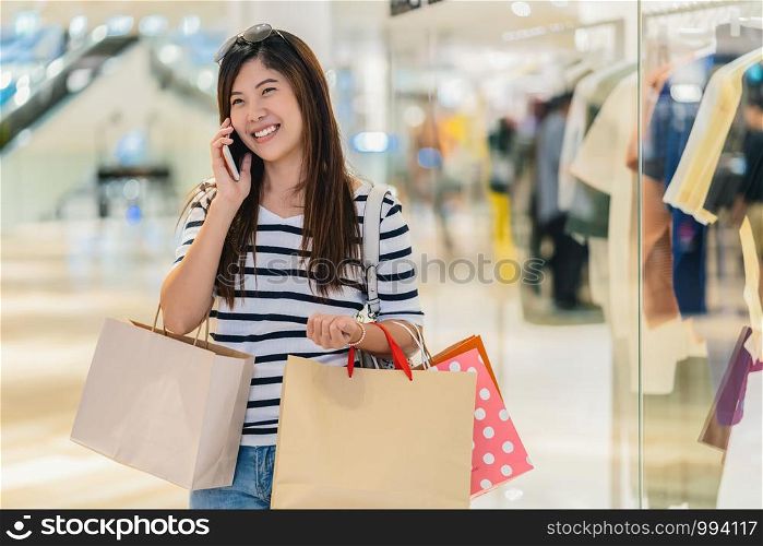 Happy Asian woman calling via the smart mobile phone for checking the product and price and holding the shopping bag beside glass shop in department store