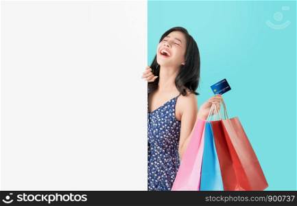 Happy Asian woman holding shopping bags with credit card and showing blank signboard on light blue background.