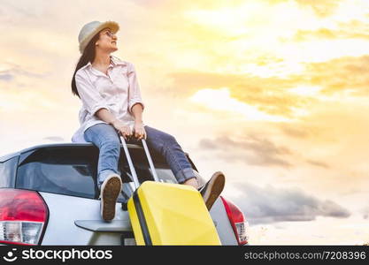 Happy Asian woman on top of car with luggage bag. Girl sitting on roof and looking sunset before night in evening. People lifestyle in long vacation trip concept. Nature and transportation vehicle.