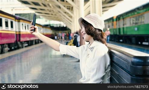 Happy Asian woman passenger with casual taking selfie by smart mobile phone in train station rails for travel in Bangkok city, Thailand. lifestyle and transportation concept.