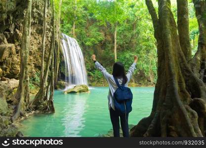 Happy Asian woman relaxing and enjoying at Erawan waterfall with trees in the tropical forest during travel trip and holidays vacation outdoors at national park, Kanchanaburi, Thailand.
