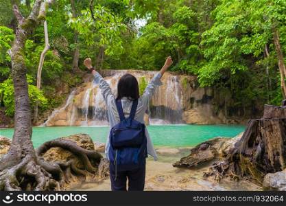 Happy Asian woman relaxing and enjoying at Erawan waterfall with trees in the tropical forest during travel trip and holidays vacation outdoors at national park, Kanchanaburi, Thailand.