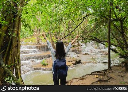 Happy Asian woman relaxing and enjoying at waterfall with trees in the tropical forest during travel trip and holidays vacation outdoors at national park, Kanchanaburi, Thailand.