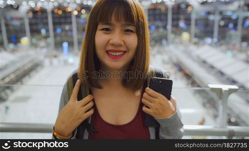 Happy Asian woman smiling to camera while staying at terminal in international airport, female tourist ready to travel on vacation. Lifestyle women happy holiday in the airport concept.