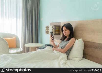 Happy Asian woman smiling, using a mobile phone, chatting with friends on bed in a modern bedroom with white blanket in the morning.