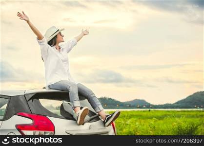 Happy Asian woman spread arms widely and breathed fresh air with happiness mood in evening after sunset on car roof. People lifestyle in long vacation trip concept. Outdoors nature and transportation