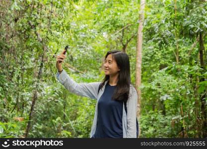 Happy Asian woman taking a selfie to post on social media at the tropical forest with trees during travel trip and holidays vacation outdoor at national park, Kanchanaburi, Thailand.