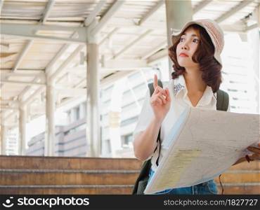 Happy Asian woman traveler tourist wanderer with trendy look searching direction on location map while traveling abroad in summer. Young female traveler with backpack and with map on street in city.