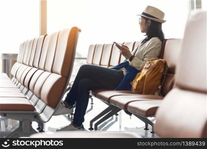 Happy Asian woman traveler wear eyeglasses and hat is enjoying with smartphone while waiting a flight at the airport on vacation. Looking away. Wireless technology.