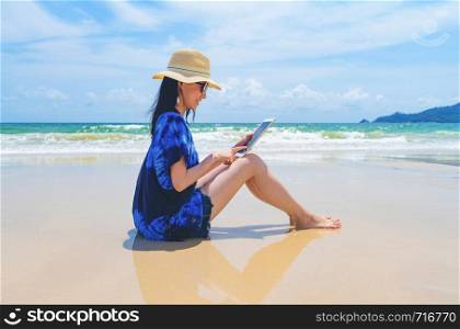 Happy Asian woman using a tablet at the beach during travel holidays vacation outdoors at ocean or nature sea at noon, Phuket, Thailand