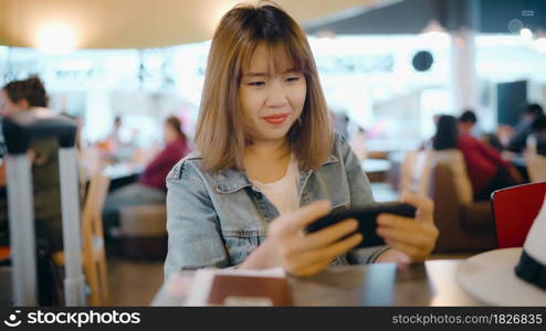 Happy Asian woman using and checking her smartphone while sitting on chair in terminal hall while waiting her flight at the departure gate in international airport. Women happy in airport concept.