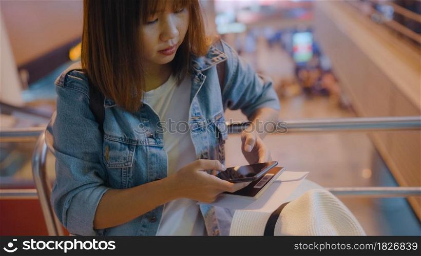 Happy Asian woman using and checking her smartphone while sitting on chair in terminal hall while waiting her flight at the departure gate in international airport. Women happy in airport concept.