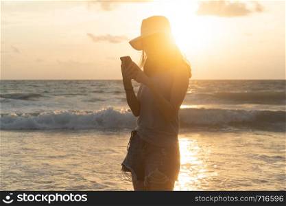 Happy Asian woman using mobile phone or texting messages on social media at beach during travel holidays vacation outdoors at ocean or nature sea at sunset time, Phuket, Thailand