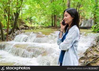Happy Asian woman using mobile smartphone to chat or call friends on social media at waterfall in the tropical forest with trees during travel trip and holidays vacation at national park, Thailand.