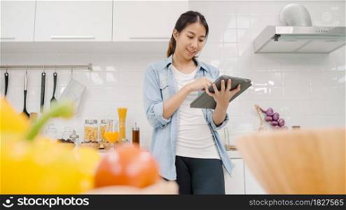 Happy Asian woman using tablet for looking recipe while making food in the kitchen, female use organic vegetable for healthy food at home. Lifestyle women making food concept.