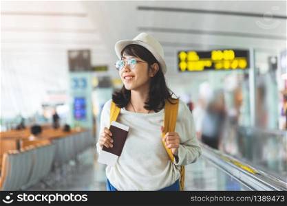 Happy Asian woman wear glasses, hat with yellow backpack is holding flying ticket, passport while waiting for the flight at the hall of airport. Smile.