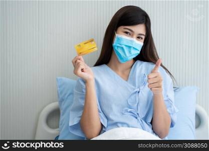 Happy Asian woman wearing a medical mask and holding mock up credit card /insurance card and ok sign in a hospital bed. Insurance policy by bank, payment medical treatment concept
