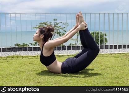 Happy Asian woman wearing black sport wear practice yoga bow or dhanurasana pose with beautiful sea,Feeling relax and comfortable,Healthy Concept