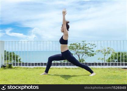 Happy Asian woman wearing black sport wear practice yoga Warrior one pose with beautiful sea view,Feeling relax and comfortable,Healthy Concept