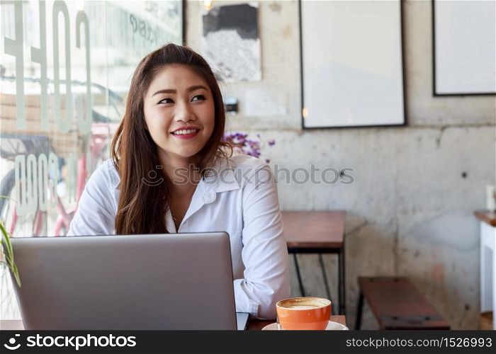 Happy Asian woman wearing white shirt smile and thinking in Coffee shop cafe with computer laptop on the table
