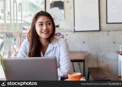 Happy Asian woman wearing white shirt smile and thinking in Coffee shop cafe with computer laptop on the table