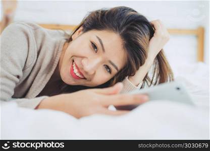 Happy Asian women are using smart phone on the bed in morning. A. Happy Asian women are using smart phone on the bed in morning. Asian woman in bed checking social apps with smartphone. Smiling woman surfing net with cellphone at home. Mobile addict concept.