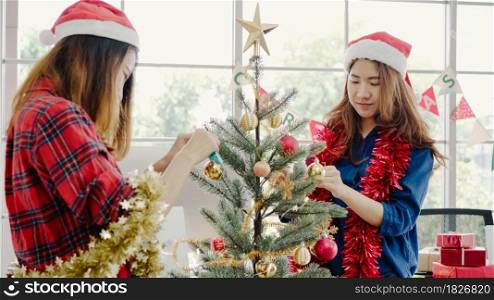 Happy Asian women holding xmas ornament for decorate christmas tree in her office. Attractive female and her friends spend holiday celebration. Winter holidays concept.