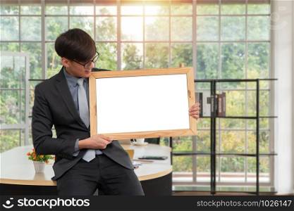 Happy Asian young businessman holding an empty white board and sitting on the table in office. Concept of copy space for advertisement, text, and content.