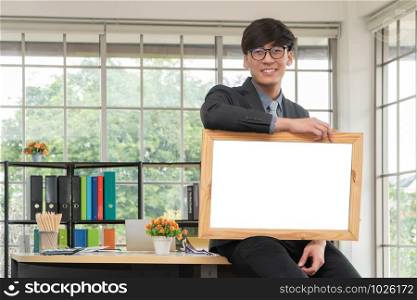 Happy Asian young businessman holding an empty white board and sitting on the table in office. Concept of copy space for advertisement, text, and content.