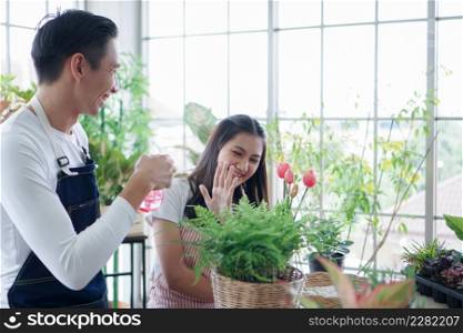 Happy Asian young couple in apron enjoys teasing each other by spraying water from a spray bottle while caring for plants in greenhouse at home
