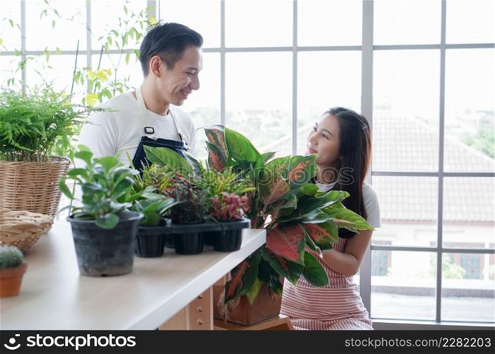 Happy Asian young couple in apron smiling at each other as they look after plants in greenhouse at home