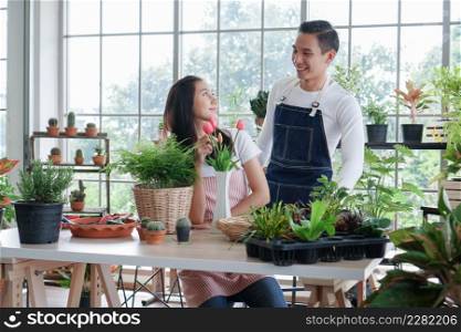 Happy Asian young couple in apron smiling at each other as they look after plants in greenhouse at home