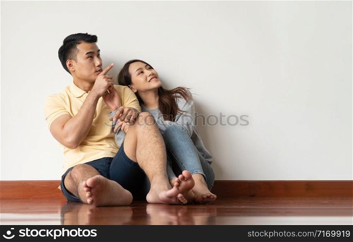 Happy Asian young couple sitting on floor at home and looking up with copy space pointing up into the air on the background of the white wall