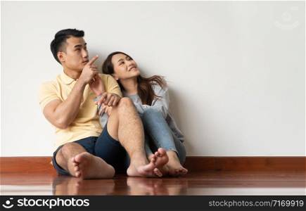 Happy Asian young couple sitting on floor at home and looking up with copy space pointing up into the air on the background of the white wall