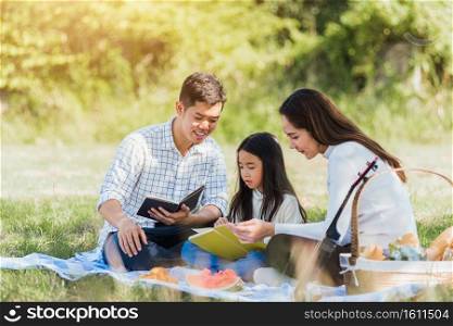 Happy Asian young family father, mother and child little girl having fun and enjoying outdoor on picnic blanket reading book in park at sunny time, summer leisure spring concept