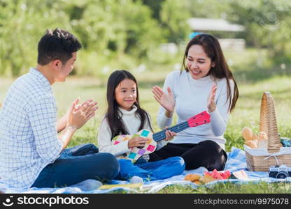 Happy Asian young family father, mother and children having fun and enjoying outdoor together sitting on the grass party with playing Ukulele during a picnic in the garden park on a sunny day