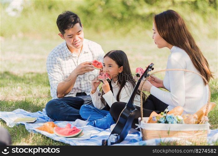 Happy Asian young family father, mother andχld litt≤girl having fun and enjoying outdoor sitting onπcnic blanket eating watermelon fruit snack lunch in park sunny time,∑mer≤isure concept