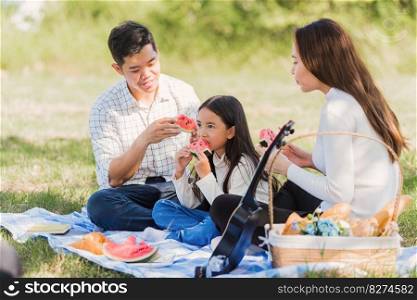 Happy Asian young family father, mother andχld litt≤girl having fun and enjoying outdoor sitting onπcnic blanket eating watermelon fruit snack lunch in park sunny time,∑mer≤isure concept
