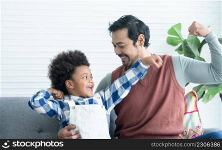 Happy Asian young father enjoy playing with little African kid boy in living room at home. Dad and son playfully show off the muscles of their arms. Diverse family concept