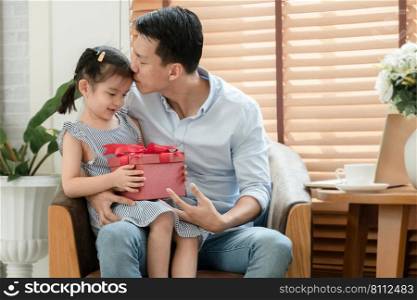 Happy Asian young father kissing forehead of his little cute daughter who surprise give him red gift box and sitting on lap while he is working with laptop in living room at home on father"s day
