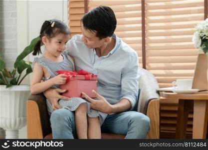 Happy Asian young father smiling and looking at little cute daughter who surprise give him red gift box and sitting on lap while he is working with laptop in living room at home on father"s day
