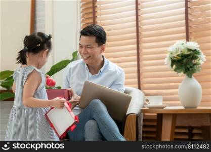 Happy Asian young father smiling with surprise face receive and open red gift box from little cute daughter while he is working with laptop in living room at home on father"s day