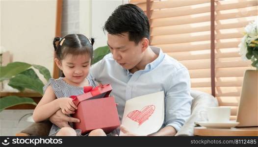 Happy Asian young father surprise with red gift box and heart colored card which little cute daughter give him and sitting on lap while he is working with laptop in living room at home on father"s day