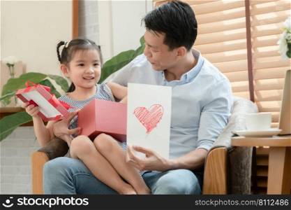 Happy Asian young father surprise with red gift box and heart colored card which little cute daughter give him and sitting on lap while he is working with laptop in living room at home on father"s day