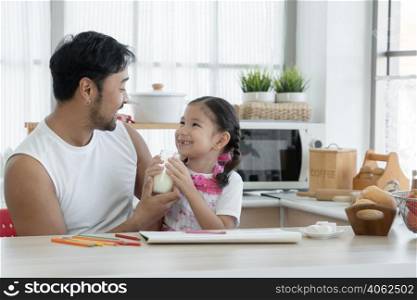 Happy Asian young father with beard holding feeding glass of milk for little cute daughter drinking while drawing with colored pencil together at home with marshmallows and breads on kitchen table