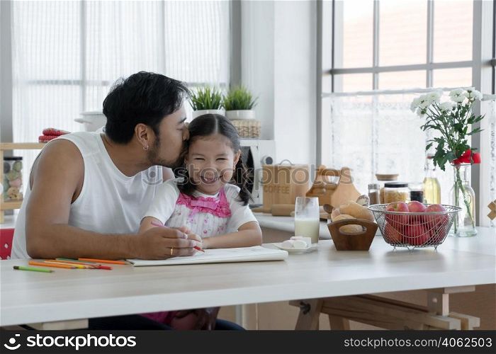 Happy Asian young father with beard kissing head of little cute daughter while drawing with colored pencil together at home with glass of milk, marshmallows, breads and apples on kitchen table