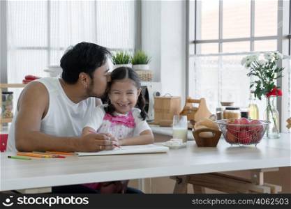 Happy Asian young father with beard kissing head of little cute daughter while drawing with colored pencil together at home with glass of milk, marshmallows, breads and apples on kitchen table